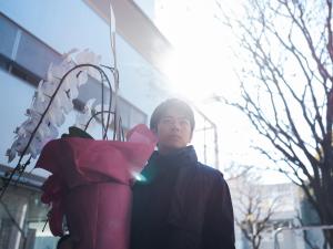 『見はらし世代』のメインビジュアル。建物の前の屋外で赤い包装が施された胡蝶蘭の鉢植えを持つ人物。